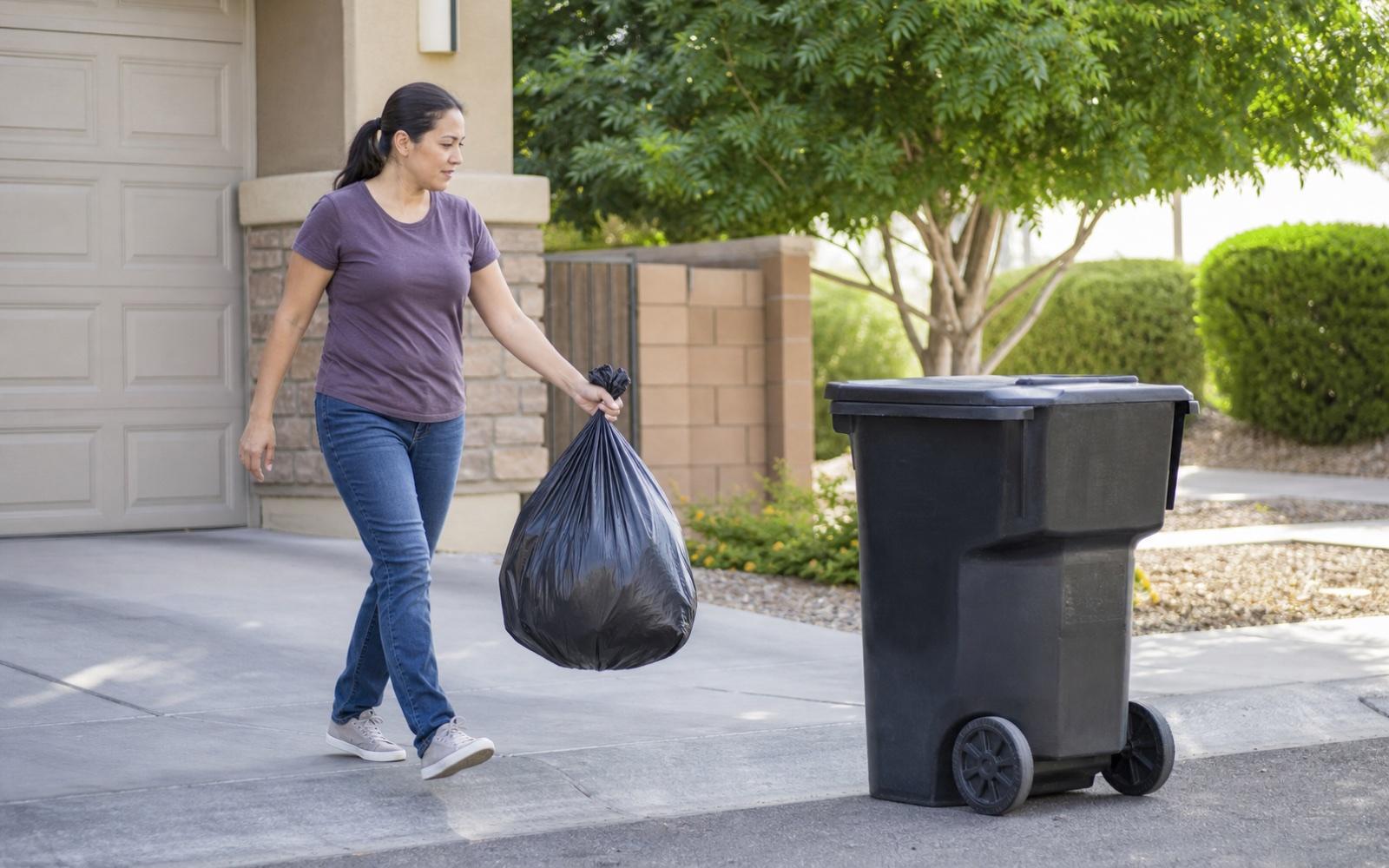 A person tying a full trash bag beside an indoor trash bin
